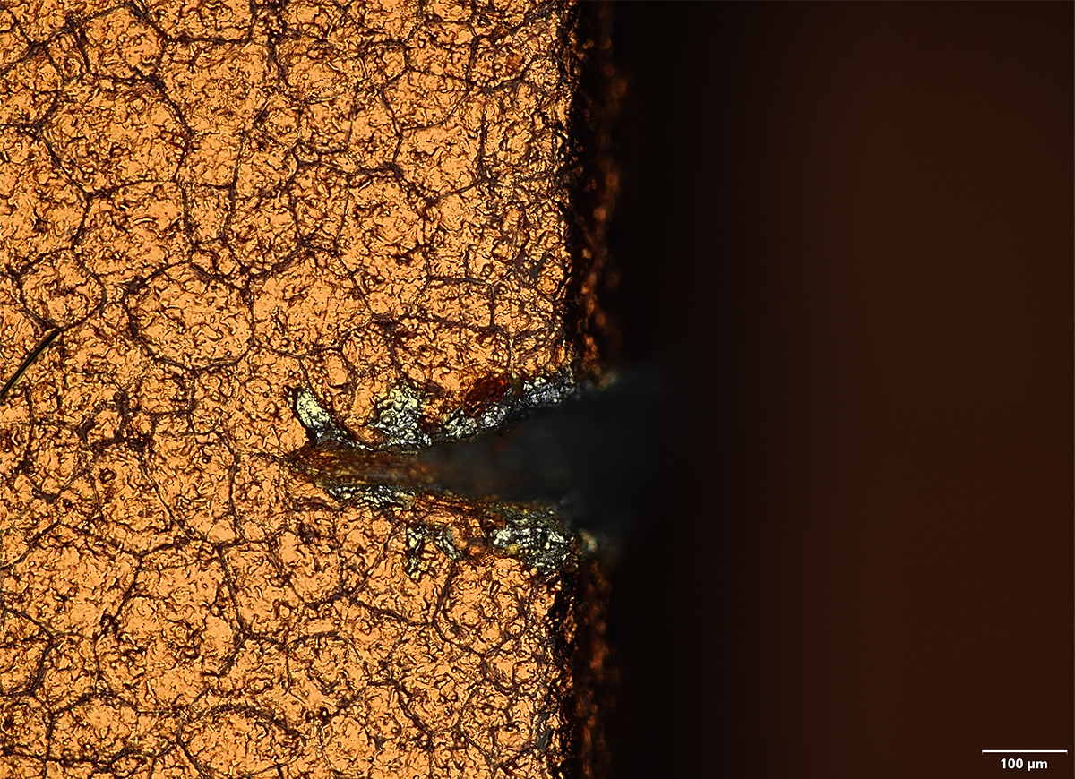 Under a Mohs level-4 pick, roughly the hardness of a coin, the anodized coating chips away at the sharp edge of the camera plateau to reveal bare aluminum. Imaged at 20X in MIX mode.